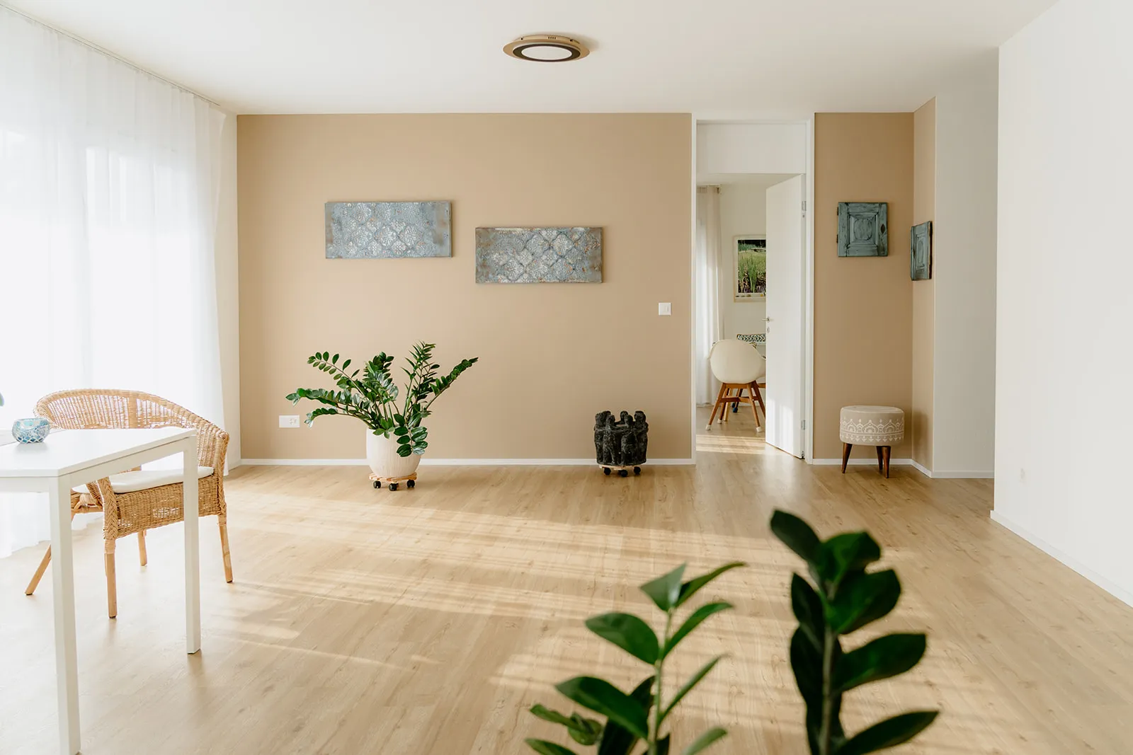 Ein heller, minimalistischer Wohnraum mit hellen Holzböden, beigen Akzentwänden, Topfpflanzen, Korbmöbeln und moderner Wandkunst bietet ein beruhigendes Ambiente - perfekt für die Praxis der Craniosacraltherapie. Das Sonnenlicht strömt durch große Fenster mit weißen Vorhängen herein.