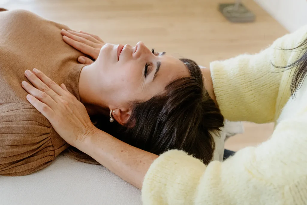 Eine Frau liegt mit geschlossenen Augen auf dem Rücken, während eine andere Person ihre Hände sanft auf ihre Brust und unter ihren Kopf legt und damit eine beruhigende Craniosacraltherapie Wil-Sitzung andeutet.