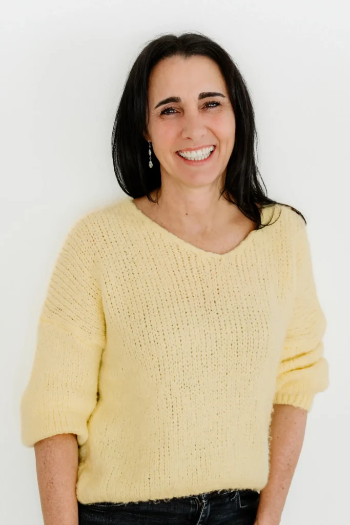 Eine Frau mit langen dunklen Haaren, die einen weichen gelben Pullover trägt, steht vor einem schlichten weißen Hintergrund und lächelt warm in die Kamera. Sie verkörpert die Ruhe und Ausgeglichenheit, die man oft nach einer Craniosacraltherapie findet.